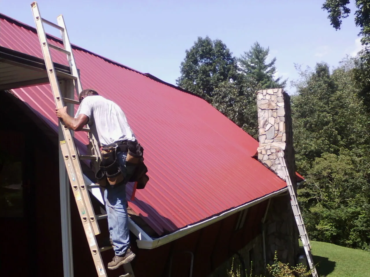 Roofer on a ladder beside a red metal roof and stone chimney — Soffit & Fascia Repair by Roofing Brothers in Haywood